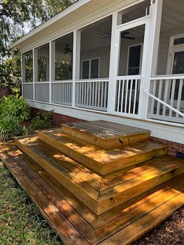 Wooden front porch stairs under a white screened porch, with railings and surrounding greenery.