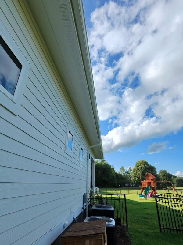 Side of a white house by a fenced backyard with a colorful playset under a cloudy blue sky