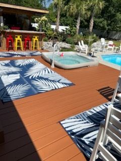 Poolside deck with blue-and-white rugs, lounge chairs, bar stools, and a hot tub in sunlight