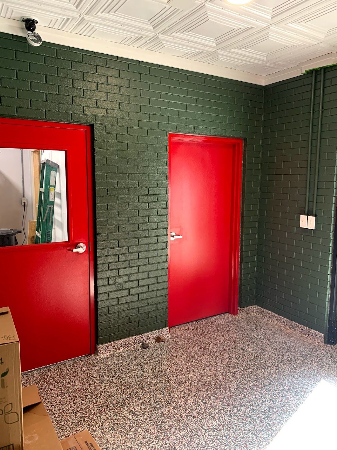 Red doors in a room with green brick walls and speckled flooring; a surveillance camera is on the ceiling.