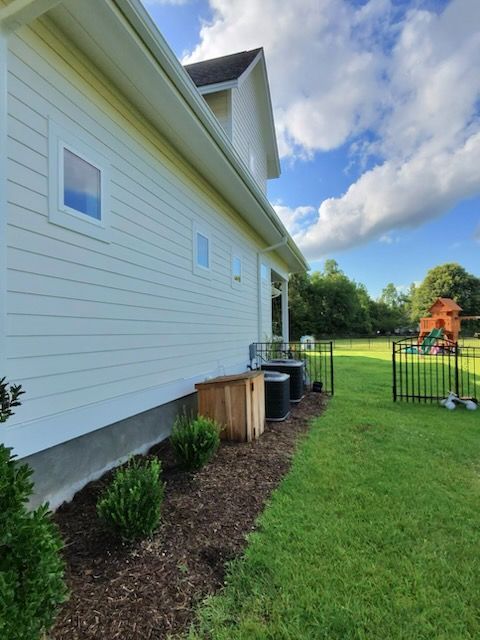 Side of a white house with mulch bed and outdoor units beside a green backyard and fence.