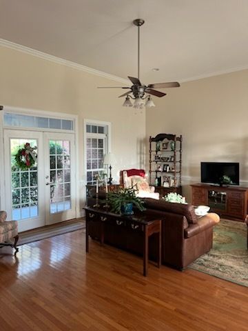 Bright living room with hardwood floors, brown sofa, TV, shelving, and French doors to a decorated patio