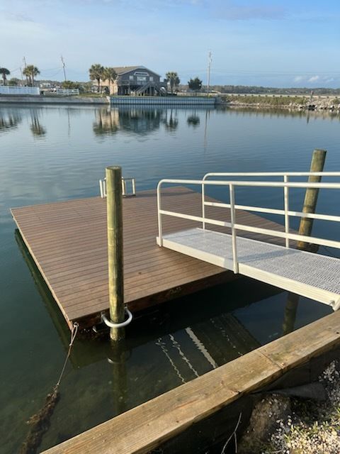Small wooden dock with white ramp over calm water, reflected buildings and palms in the background