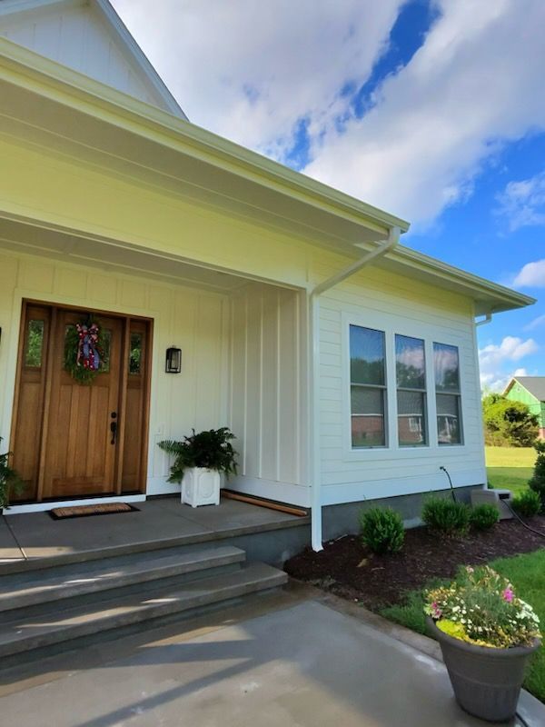Front porch of a white house with wooden door, steps, windows, and potted plants under a blue sky.