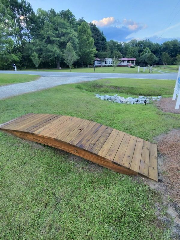 Curved wooden ramp on grass beside a driveway, with trees and a house in the background.