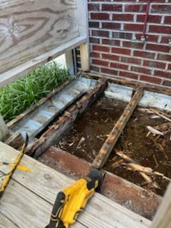 Deck boards removed, exposing wet joists and a hole beside a brick wall, with a yellow drill in the foreground