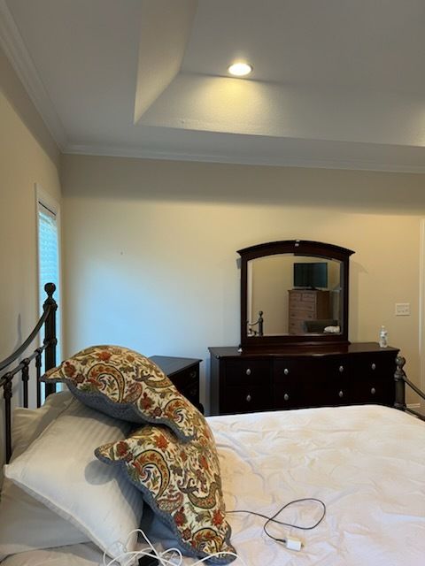 Bedroom with metal bed, patterned pillows, white bedding, and dark dresser with mirror under a tray ceiling.