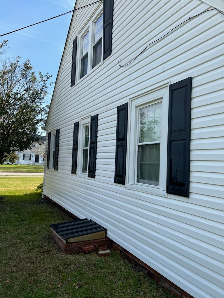 White house side with black shutters, windows, and a small basement entry over grass.