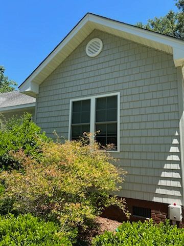 Side view of a gray house with white trim, a round attic vent, and shrubs in front under a blue sky