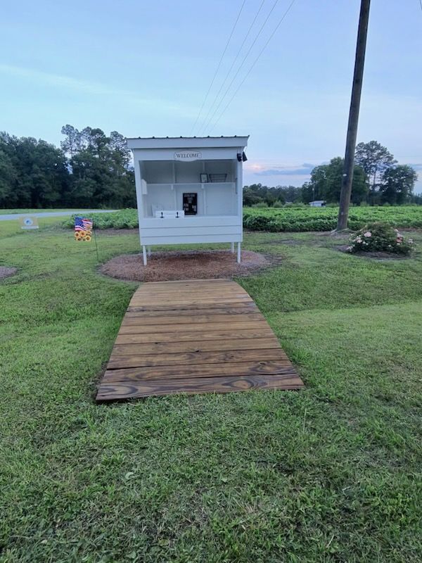 Small white shed at the end of a wooden walkway in a grassy field near trees and a utility pole.