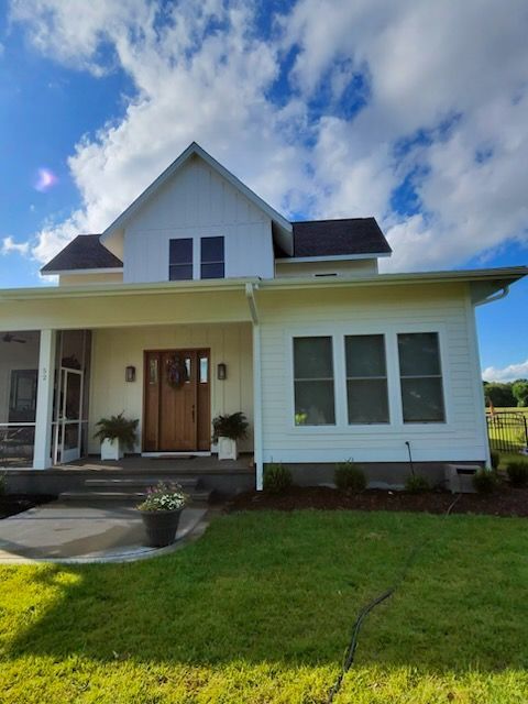 White house with a front porch, dormer windows, and a green lawn under a partly cloudy sky