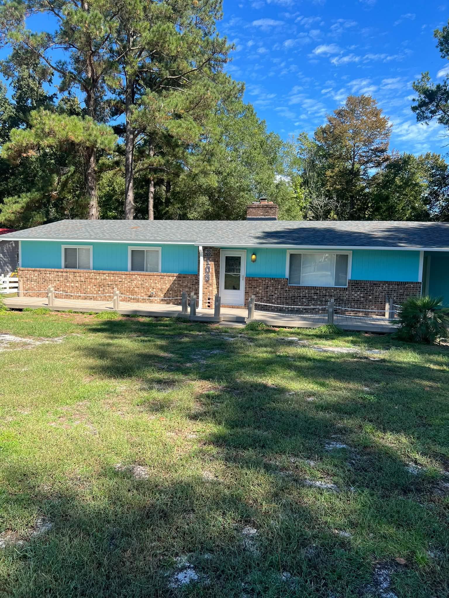 Single-story house with teal shutters, brick porch, and a grassy front yard under a blue sky