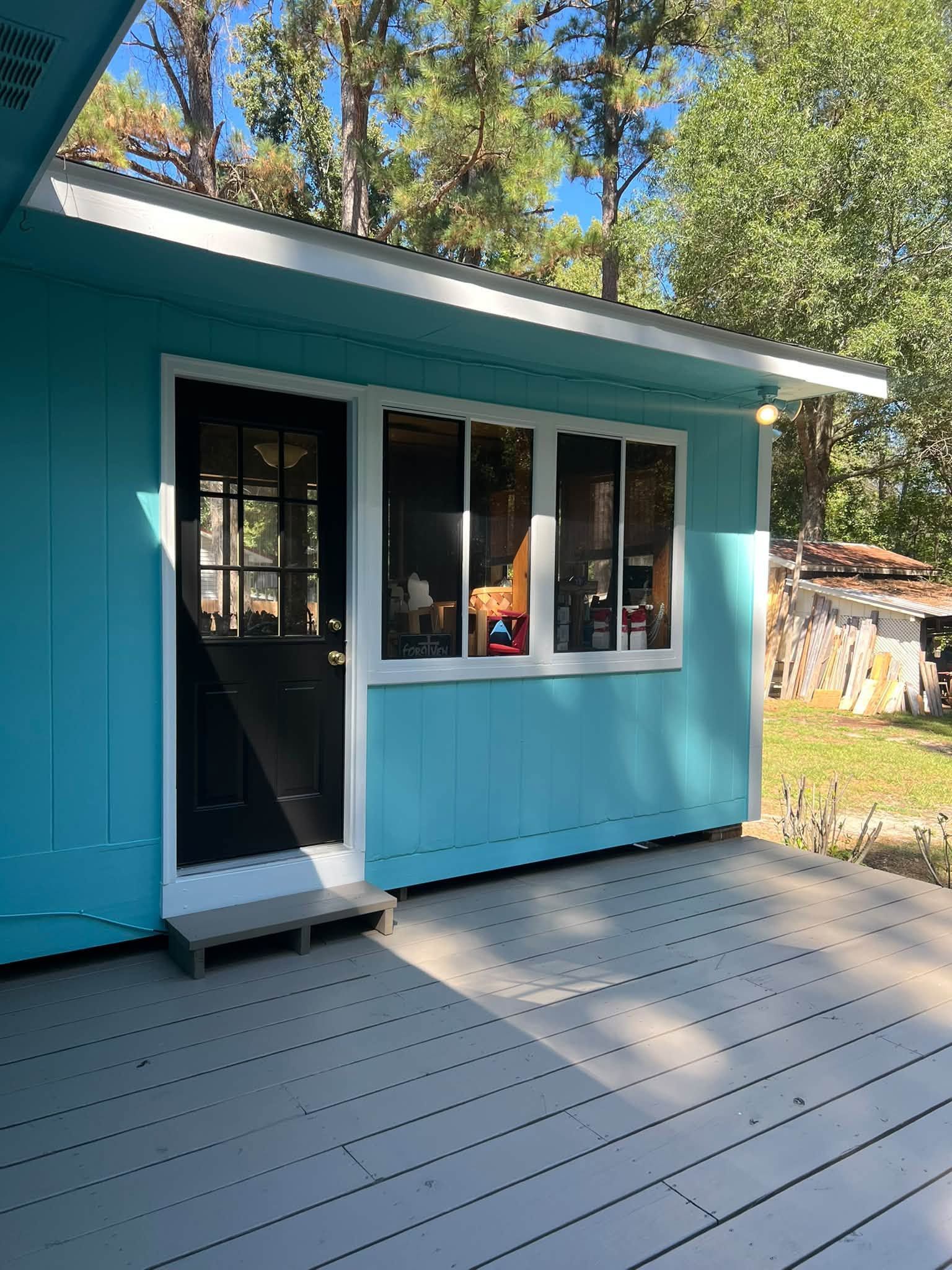 Turquoise house exterior with white-trimmed windows and a sunlit gray deck under trees