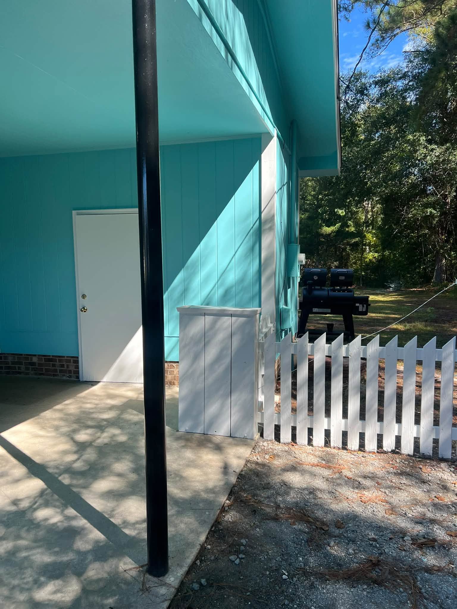 Turquoise house exterior with a white picket fence and a black support post in sunlight.