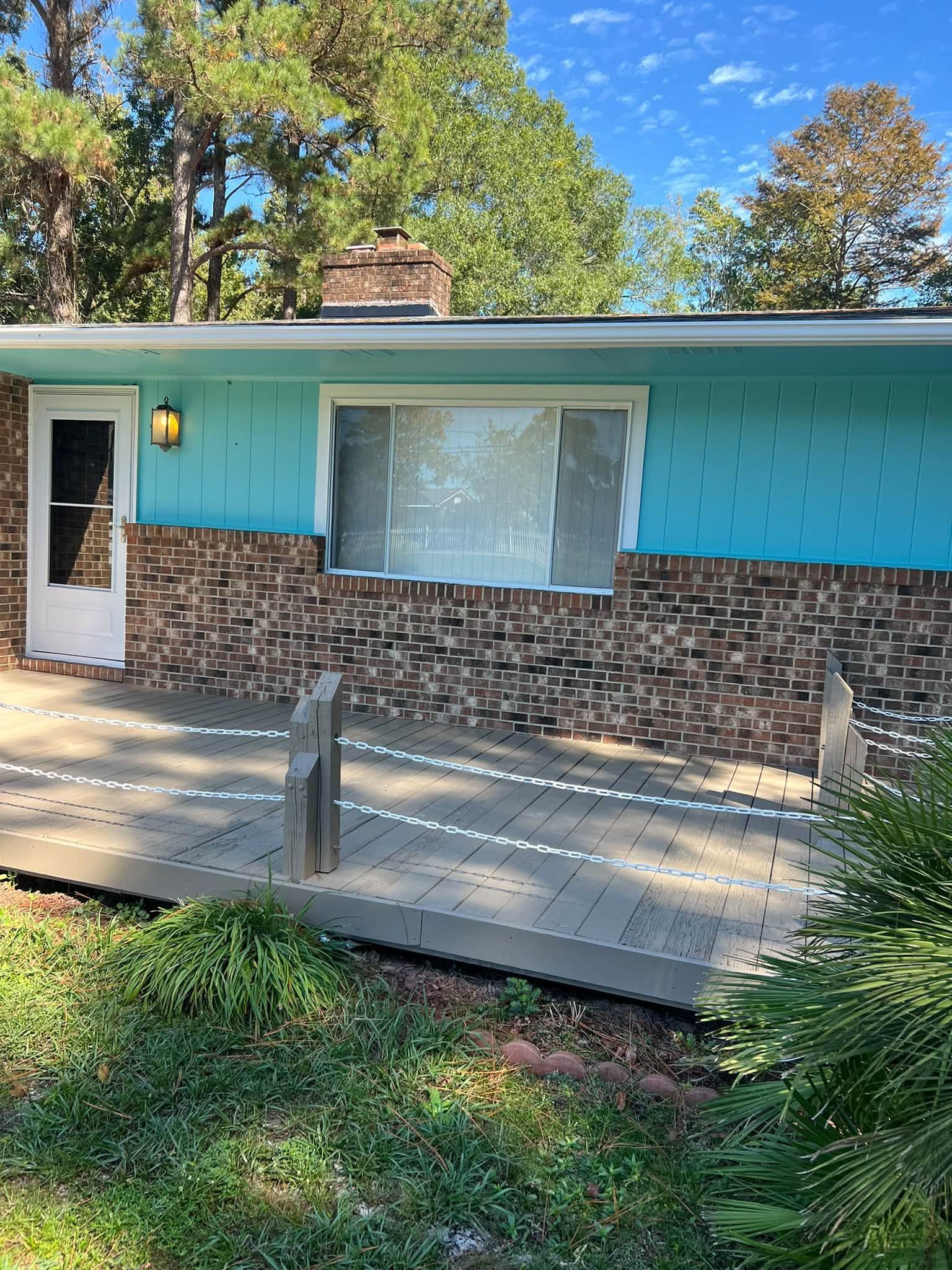 Single-story house with turquoise siding, brick base, white door, and front porch in a leafy yard