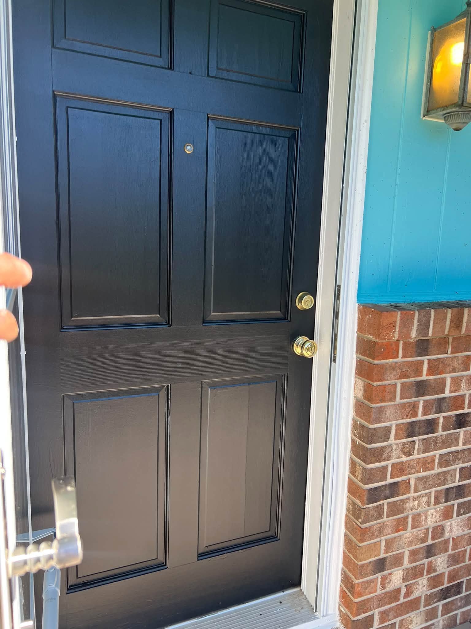 Black front door with gold hardware beside a brick wall and blue exterior siding.