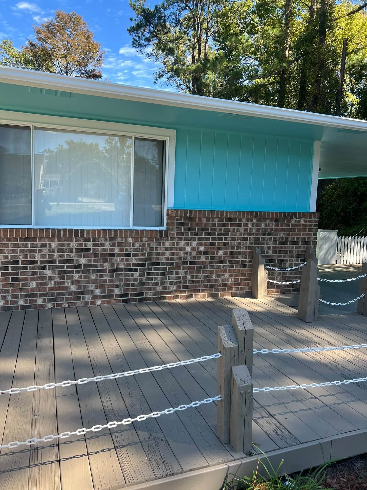 Front porch with turquoise siding, brick wall, large window, and wooden deck under trees.