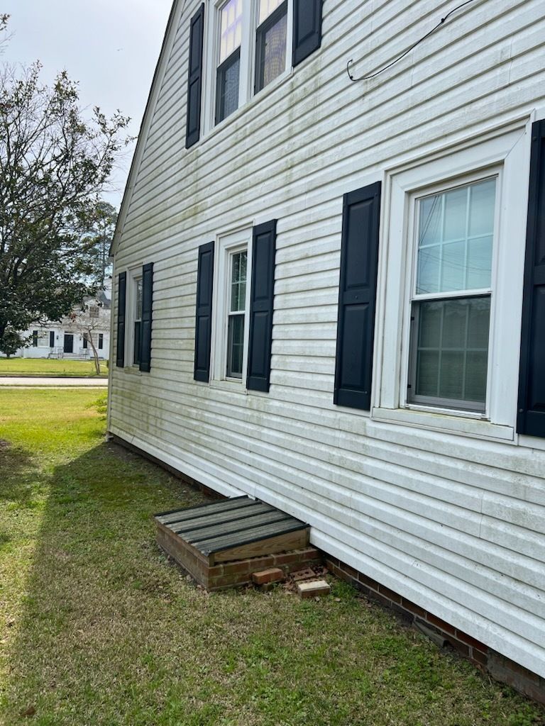 Side of a white house with black shutters and a small wooden step in the grass beneath a window.