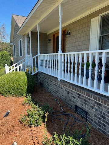 Front porch of a house with white railing, brick foundation, and landscaped shrubs along the walkway.