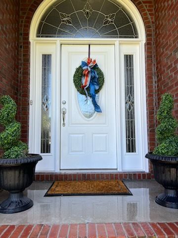 Front door with white double doors, blue holiday wreath, brick entry, and potted topiary on both sides.