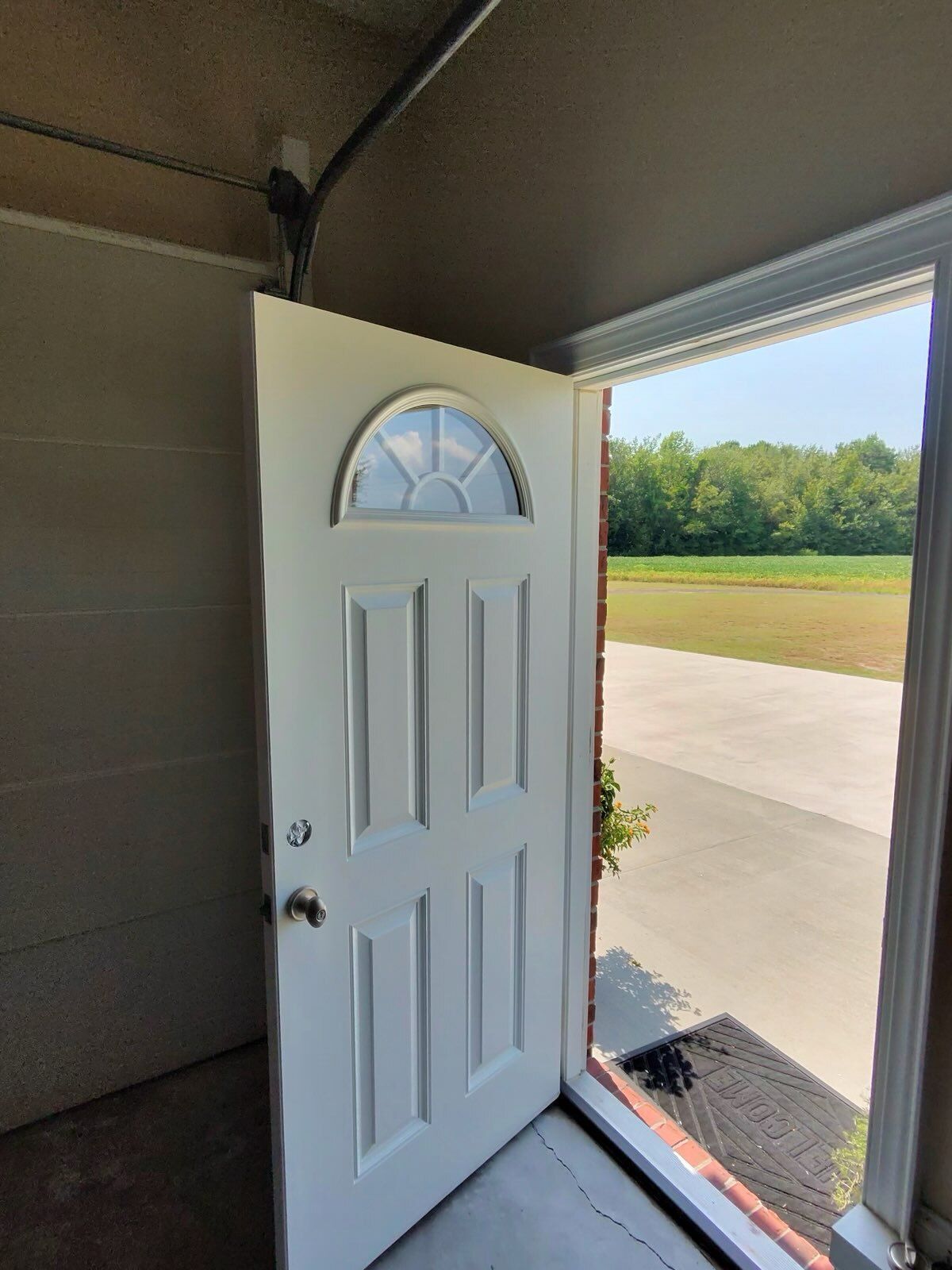 Open white front door with arched window leading to a sunlit driveway and lawn