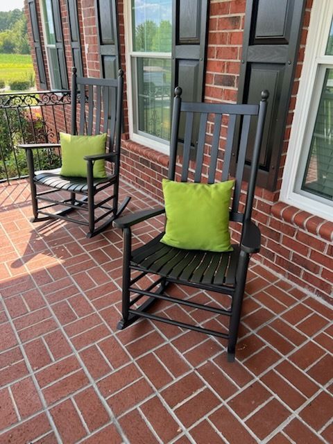 Two black rocking chairs with green cushions on a brick porch beside large windows