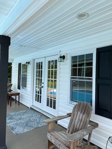 White porch with black door, French doors, patterned rug, and wooden chair beside a swimming pool.