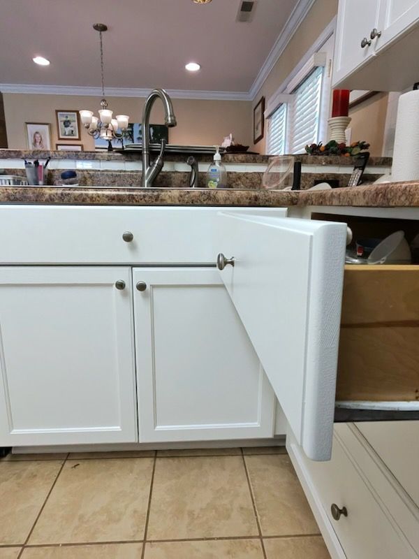Open kitchen cabinet under granite countertop, showing white cabinet doors and beige tile floor.