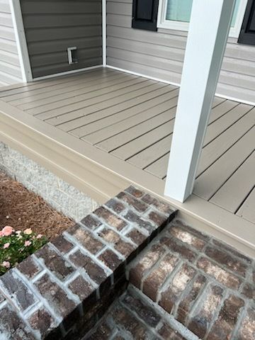 Front porch deck with beige flooring, white post, gray siding, and brick steps leading down to a garden bed