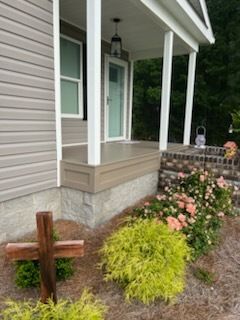 Front porch with white siding, brick steps, and a small wooden cross in a landscaped garden.
