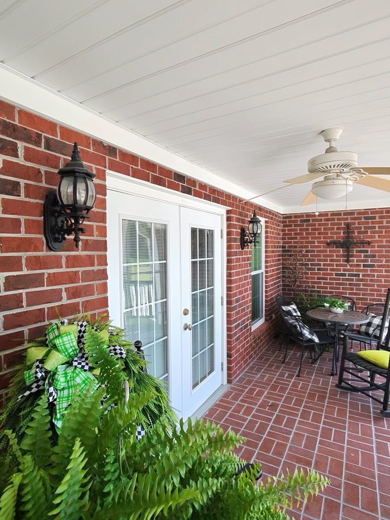 Brick porch with white French doors, hanging lanterns, ferns, and patio chairs under a ceiling fan.