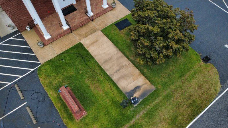 Aerial view of a sidewalk, lawn, bench, tree, and brick building beside a parking lot.