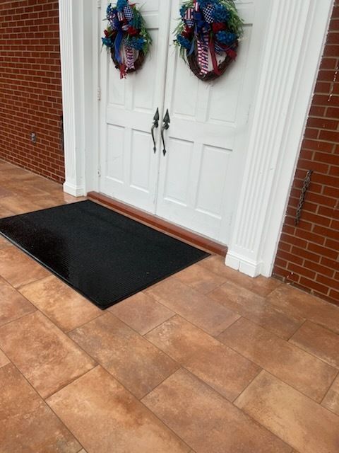 White double front doors with wreaths and a black doormat on a brick porch