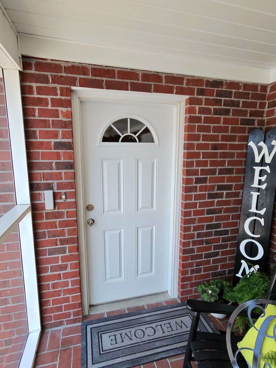 White front door with arched window on a brick porch, welcome mat, and black welcome sign.