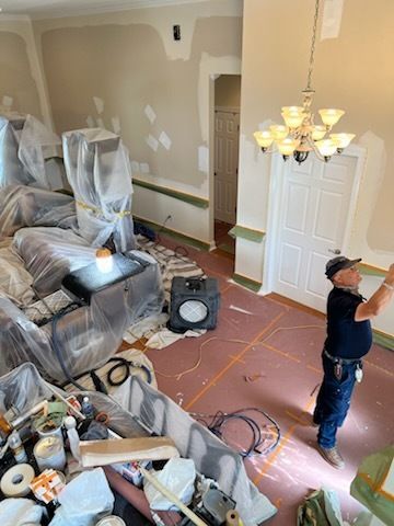 Living room under renovation with furniture covered in plastic and a worker measuring the floor.