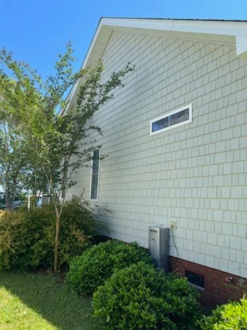 Side view of a white house with a small window, brick foundation, and shrubs beside a green lawn under blue sky