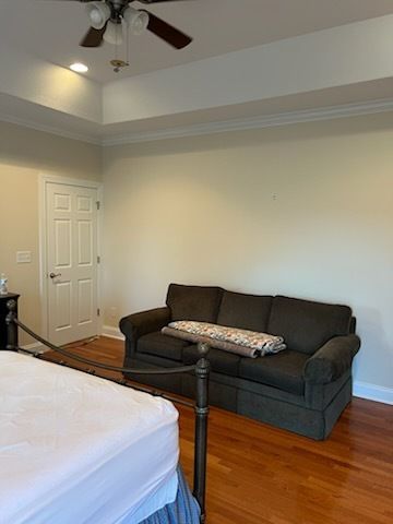 Bedroom with dark sofa, bed, wooden floor, and ceiling fan in a light-colored room