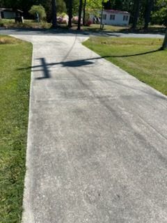 Concrete driveway leading to a road, with grass on both sides and a house in the distance.