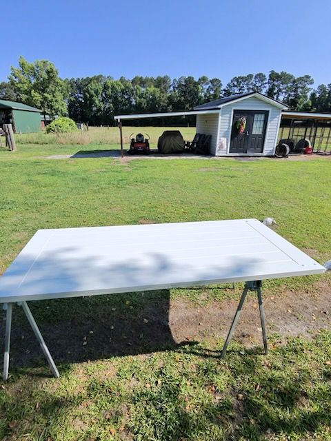 White folding table on grass in a sunny backyard with a small house and carport in the background