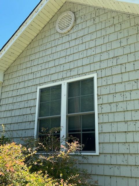 Side of a gray house with white trim, a round attic vent, and a window above shrubs.