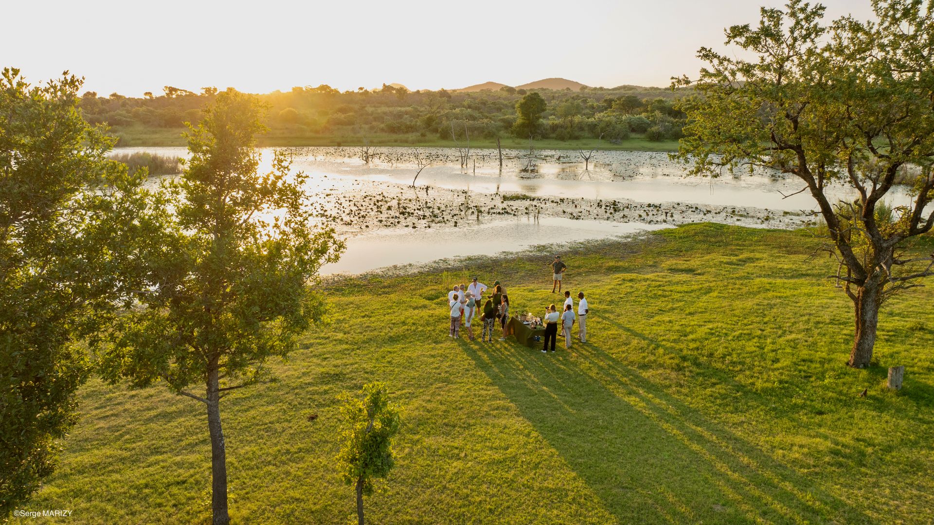 People gathered on a grassy lakeshore at sunset with trees and water in the background.
