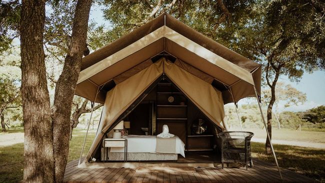 Canvas glamping tent with open front, bed inside, on a wooden deck in a sunny wooded area