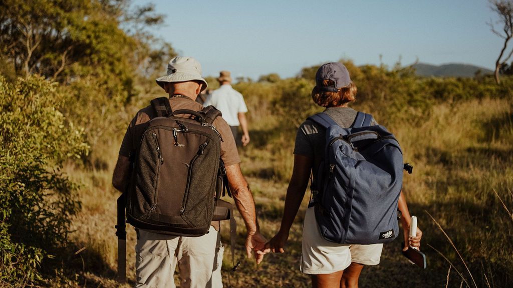 Three hikers with backpacks walking along a sunny dirt trail through brush