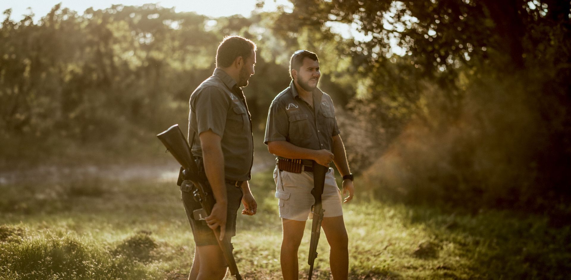 Two people standing outdoors in a sunlit field, holding rifles and looking off to the right.