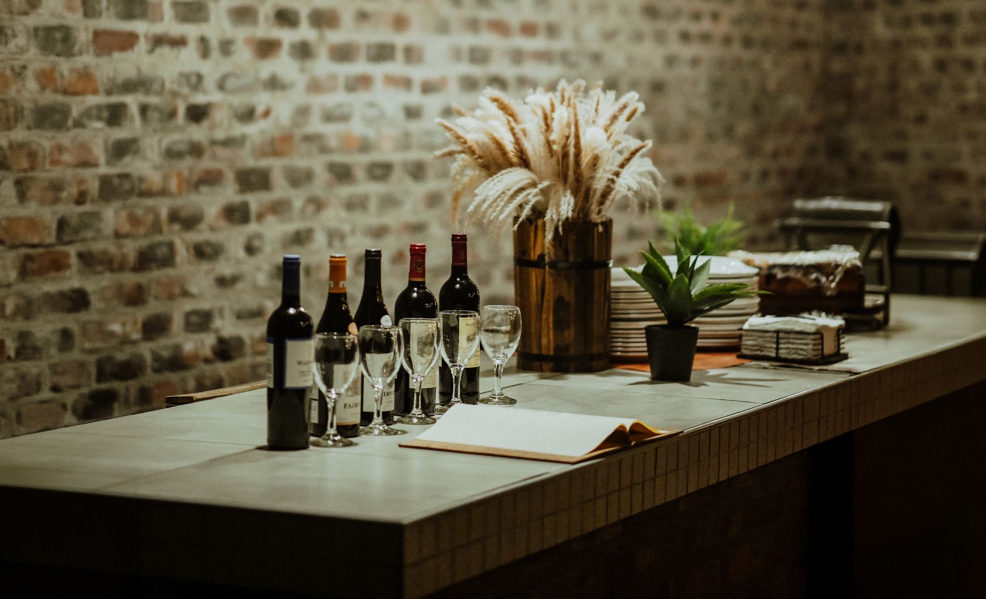 Wine bottles and a small plant arranged on a bar counter under woven hanging lights in a rustic room