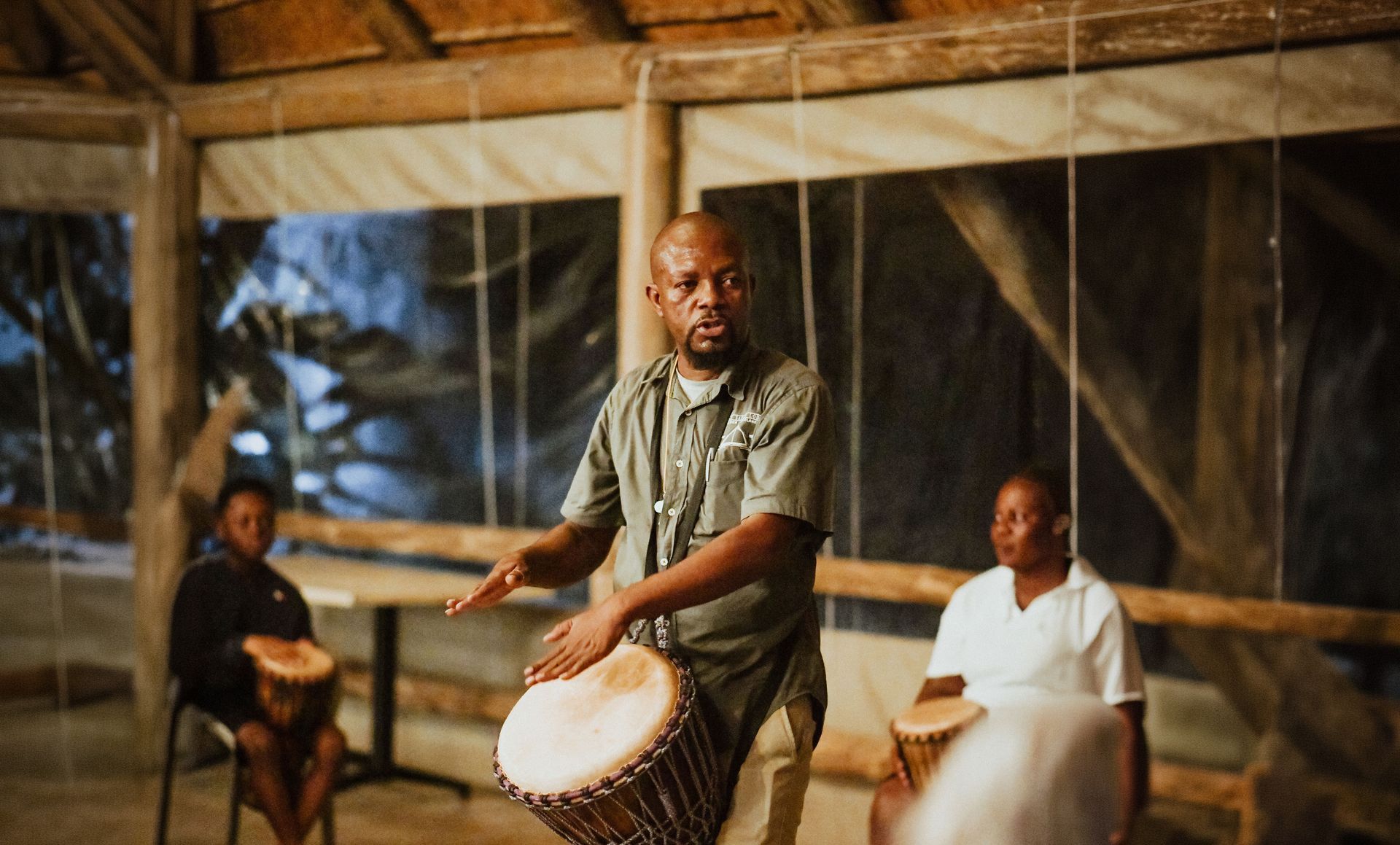 Man playing a drum in a warmly lit indoor drumming circle, with seated drummers in the background