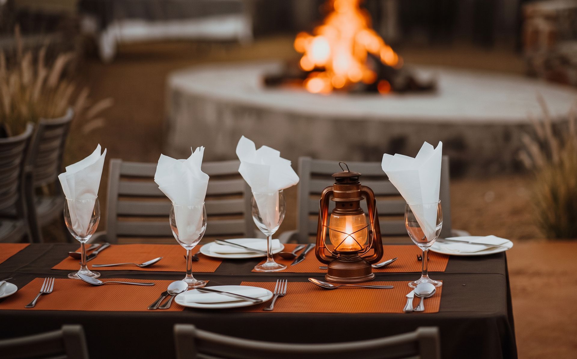 Lantern-lit outdoor dining table set for four with folded napkins, in front of a fire pit