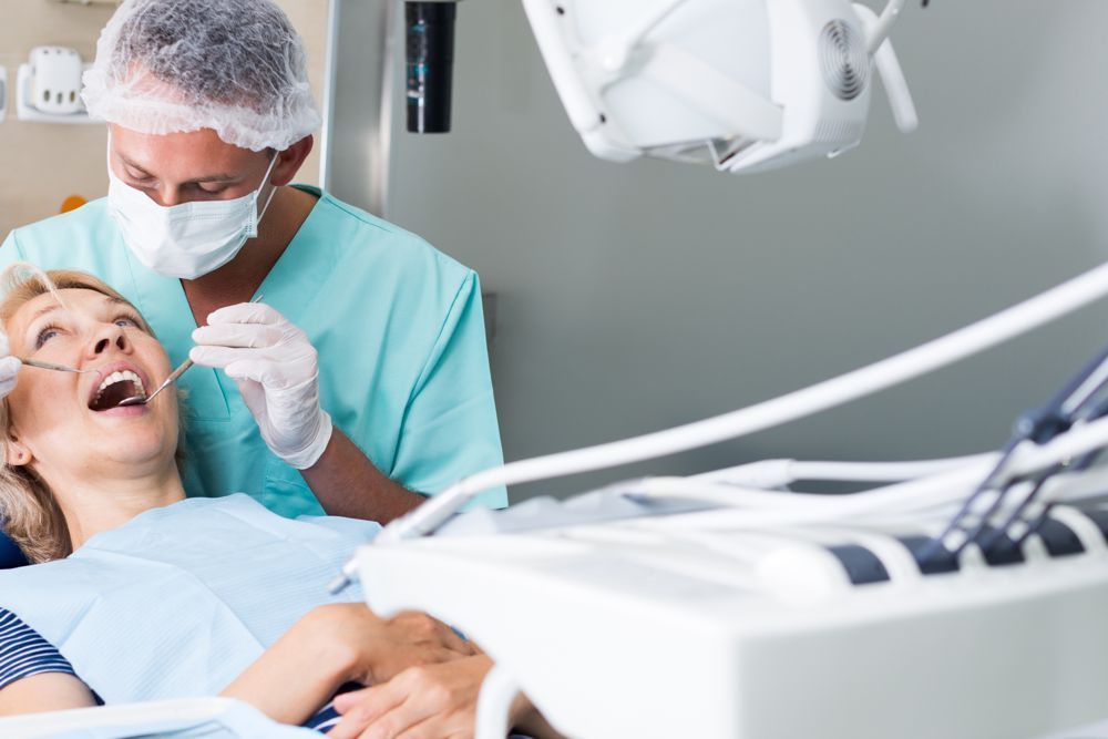 A dentist in surgical attire and a face mask examines a patient's teeth in a clinical setting.