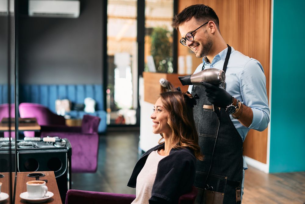 A hairstylist wearing black gloves smiles while blow-drying a client's hair in a modern salon with purple chairs.