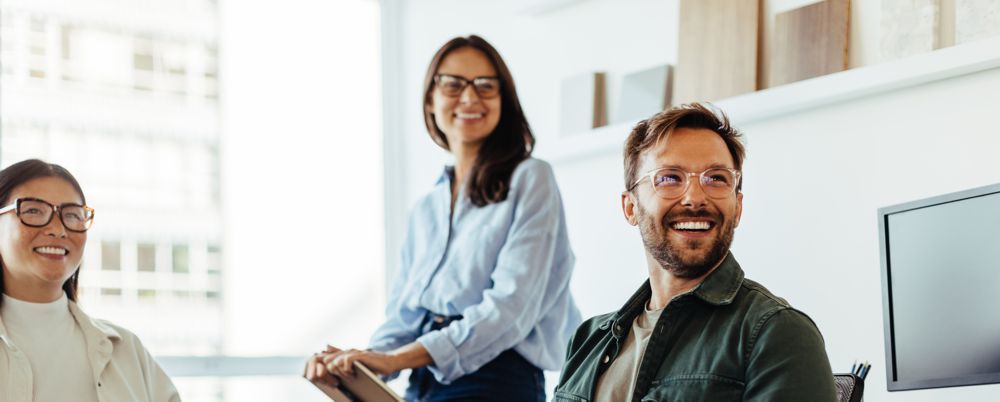 Three professionals smile while collaborating in a brightly lit, modern office workspace.